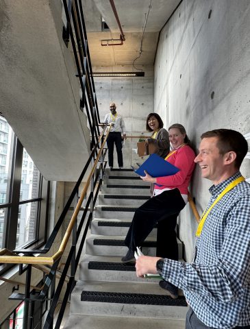 Four people stood on a staircase smiling at the camera holding notebooks to record measurements whilst auditing